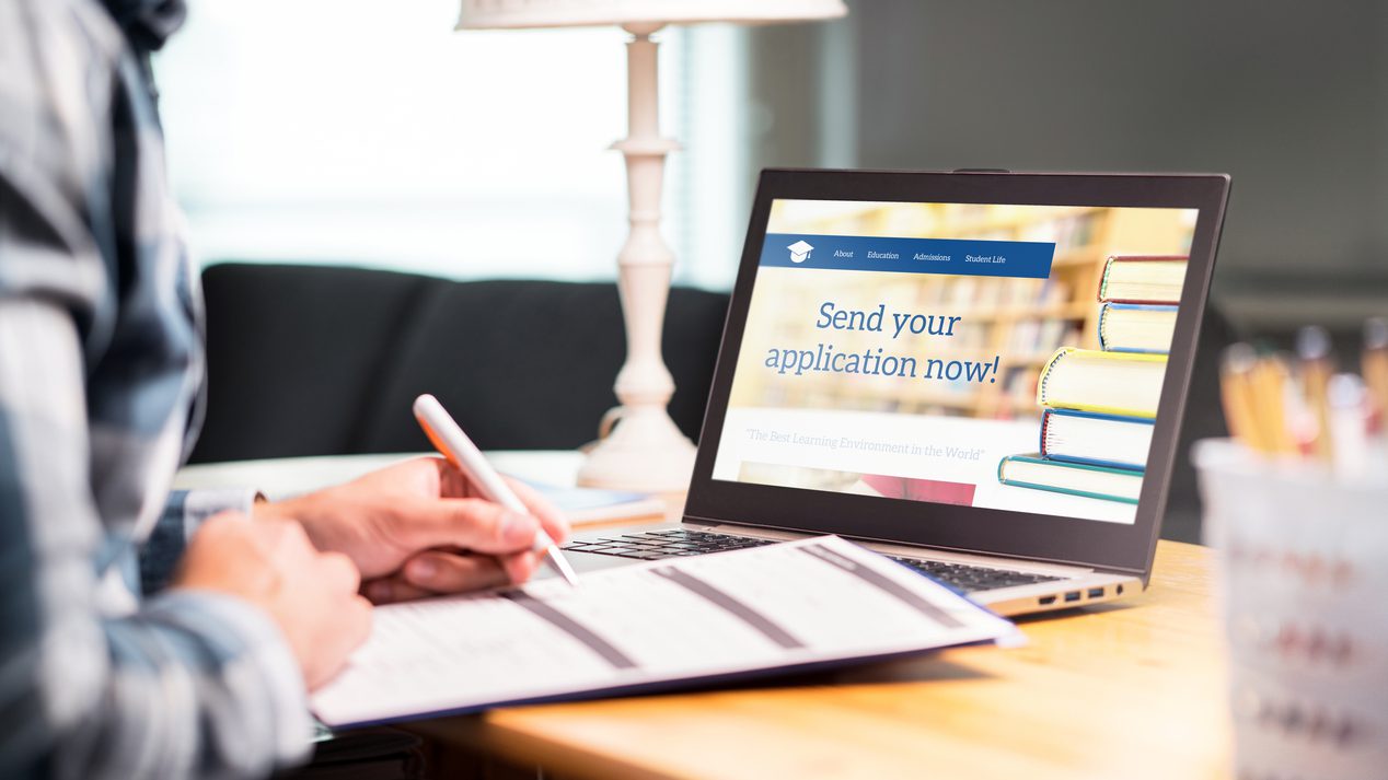 Young man writing college or university application form with pen and paper. Student applying or filling document. Scholarship, grant or admission. School website in laptop.