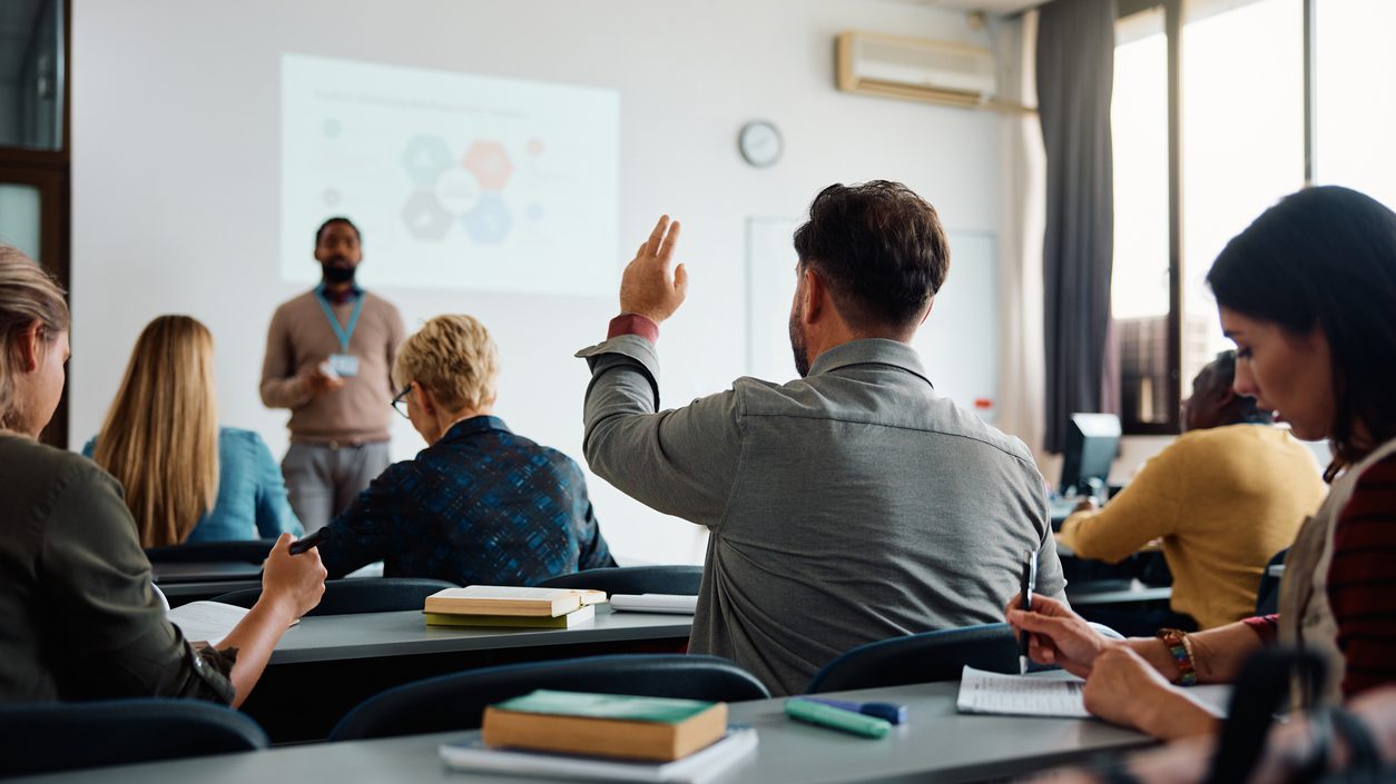 Rear view of man raising arm to ask a question during a presentation in lecture hall.